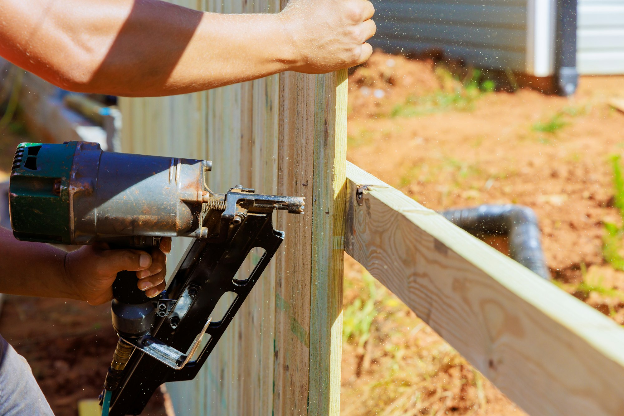 Using an air nail gun, a man constructs portions of wooden fence around his yard